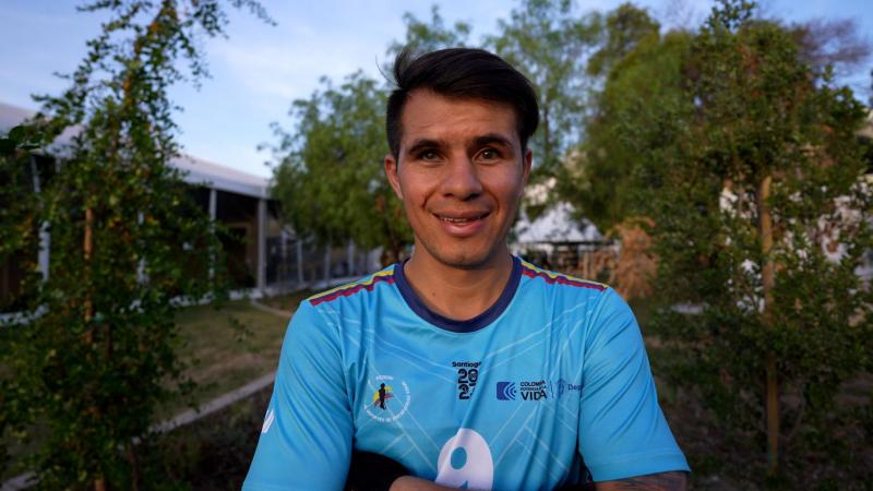 Jhon Gonzalez, a blind football player from Colombia, is standing outdoors wearing a blue uniform, crossing his arms