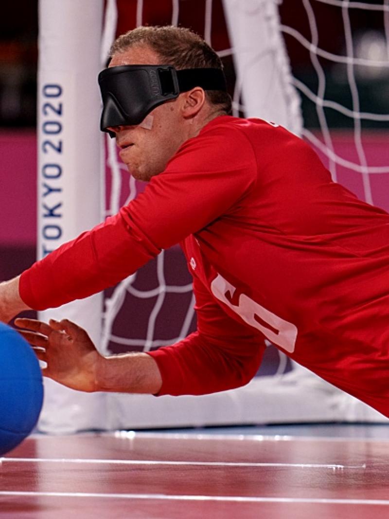 Two male goalball athletes are blocking the ball in front of a net