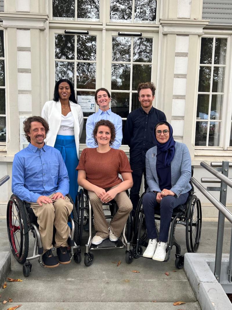 Six athletes, three standing in the back and three in wheelchairs at the front, pose for a photo in front of the IPC headquarters.