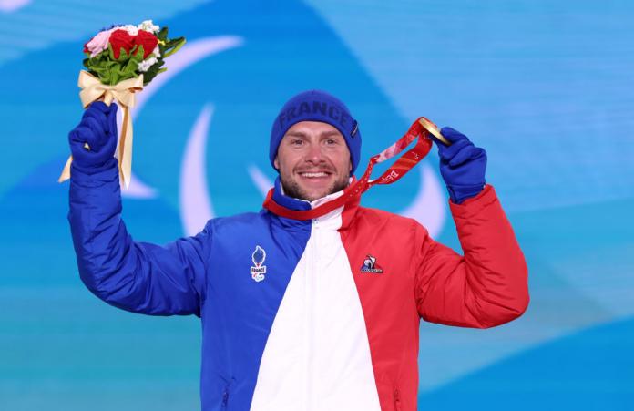 A male athlete wearing a blue, red, and white jacket and a blue cap poses for a photo on the podium. He holds a gold medal and flowers
