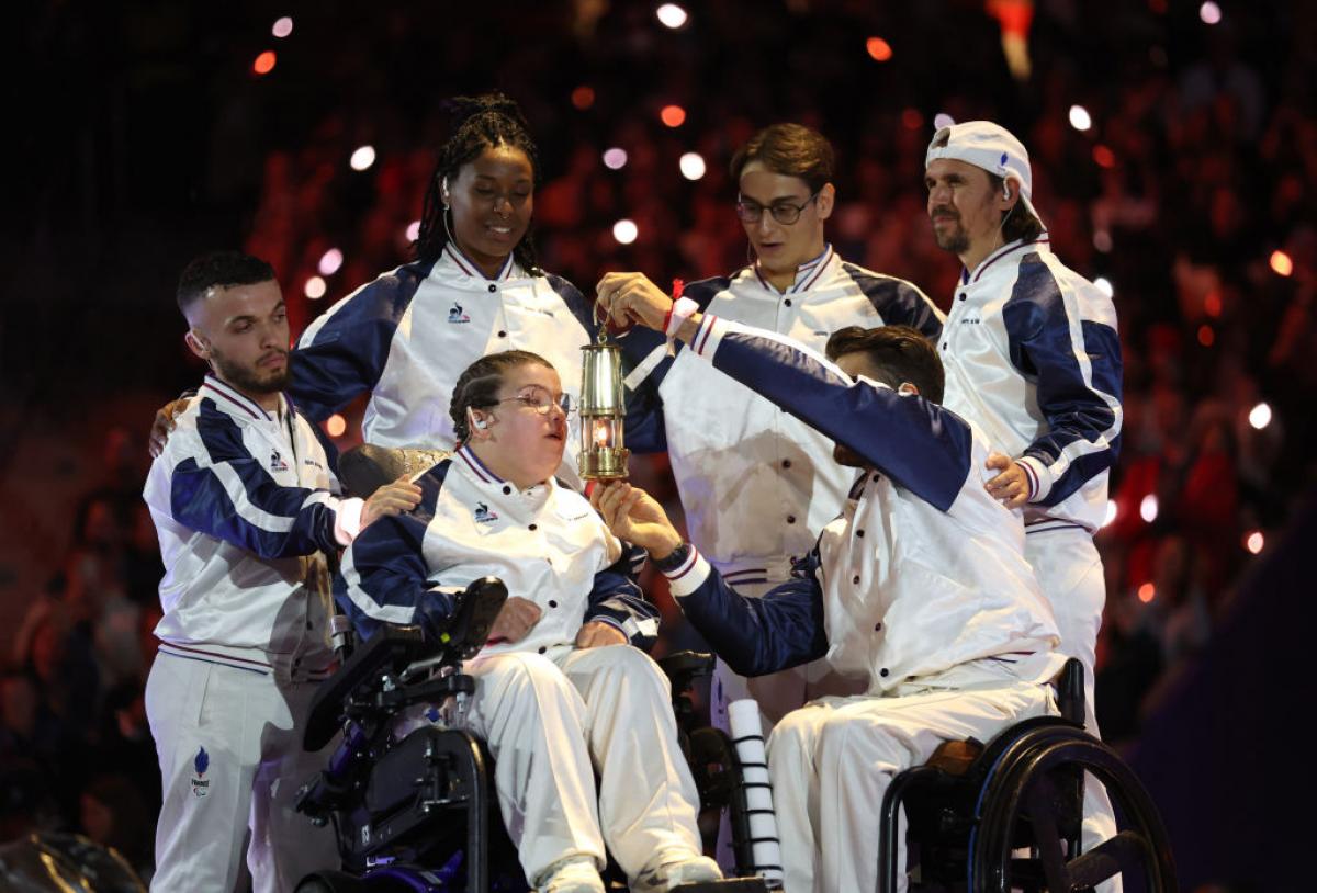 Aurelie Aubert blows out the Paralympic flame. Five athletes surround her, one of them holding a small lantern in front of her face