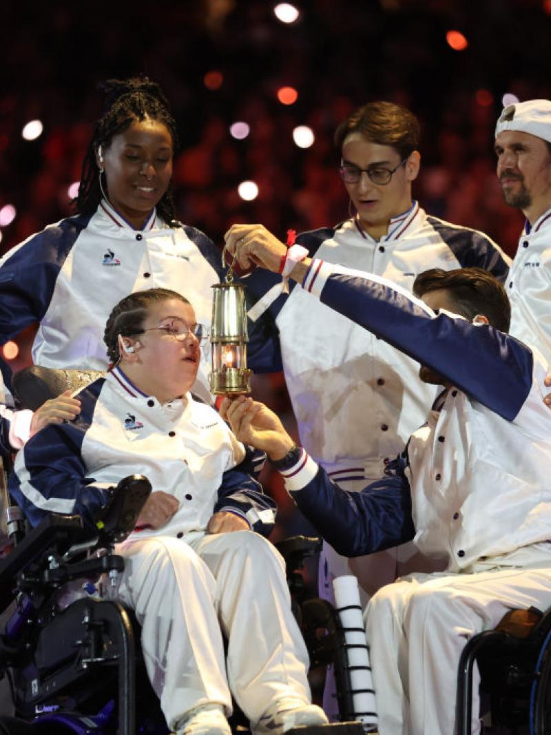 Aurelie Aubert blows out the Paralympic flame. Five athletes surround her, one of them holding a small lantern in front of her face