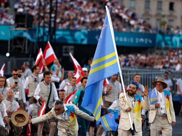A male athlete with blue hair and blue sunglasses carries Aruba's flag during the Opening Ceremony. 