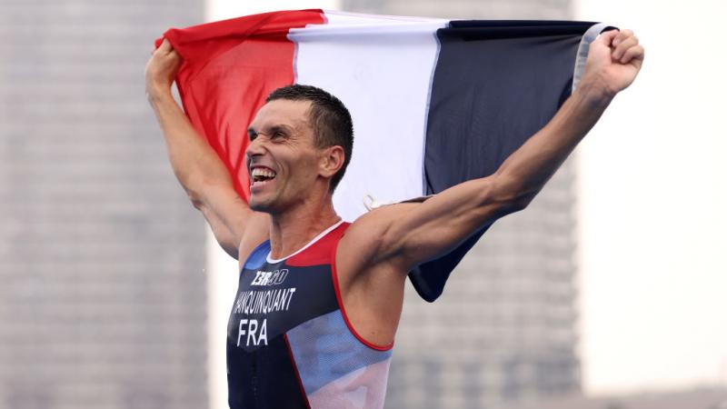 A man in a Team France triathlon uniform holds up the French flag and yells