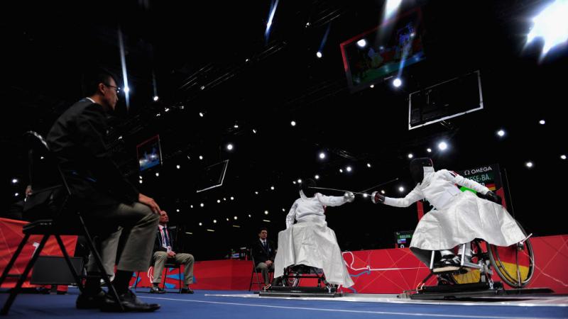 Two athletes in wheelchairs fence. They wear white uniforms and black face shields, their swords are crossed.