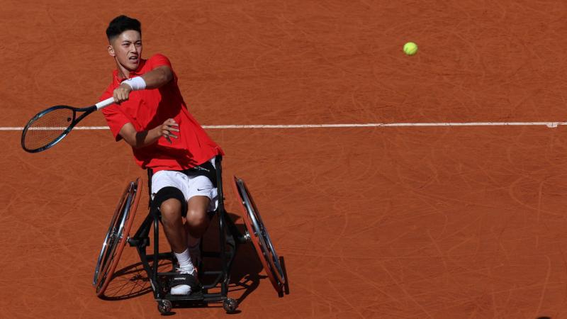 Against an orange earth background a man in a red tee and a wheelchair reaches a racket out as a yellow tennis ball flies