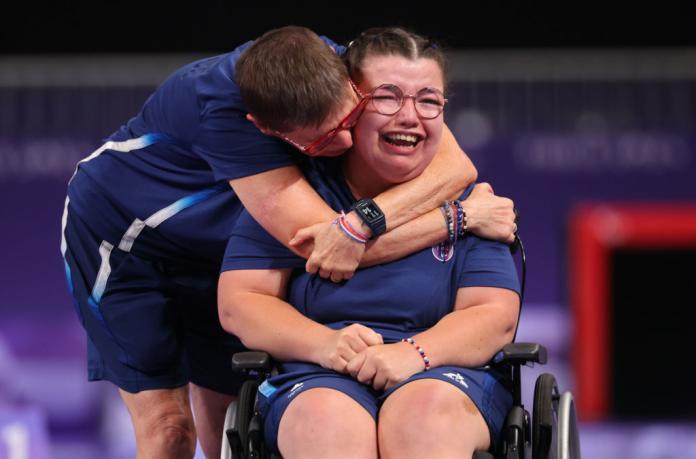 A woman in blue tee and shorts seated in wheelchair is hugged by another woman while crying