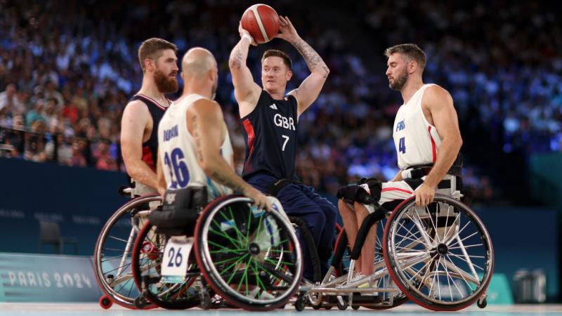A man in a wheelchair holds a basketball above his head, surrounded by three other wheelchair basketball players