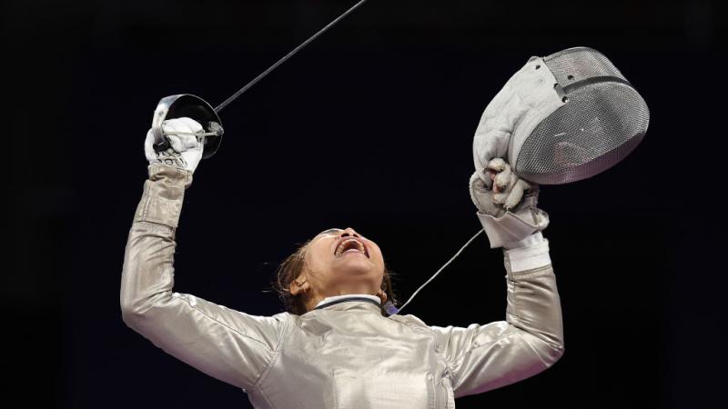 A woman competing in wheelchair fencing holds up her sabre in one hand and her face shield in the other and yells passionately