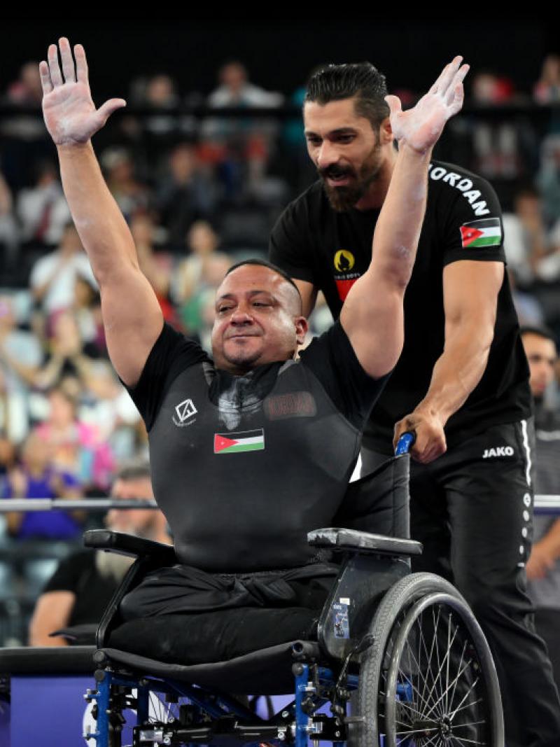 Man in black Jordan kit in wheelchair raises both hands in the air in a sign of celebration while pushed by another man with a beard
