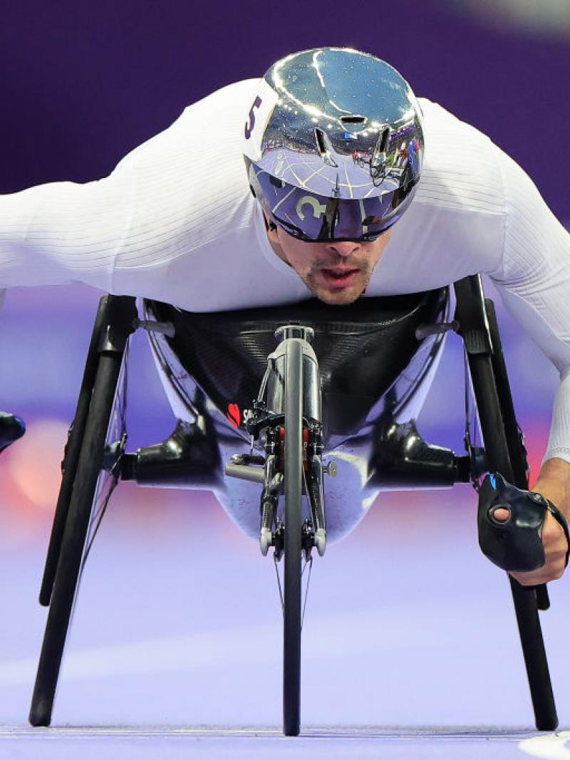 A man in a racing wheelchair and a silver helmet competes on a purple track