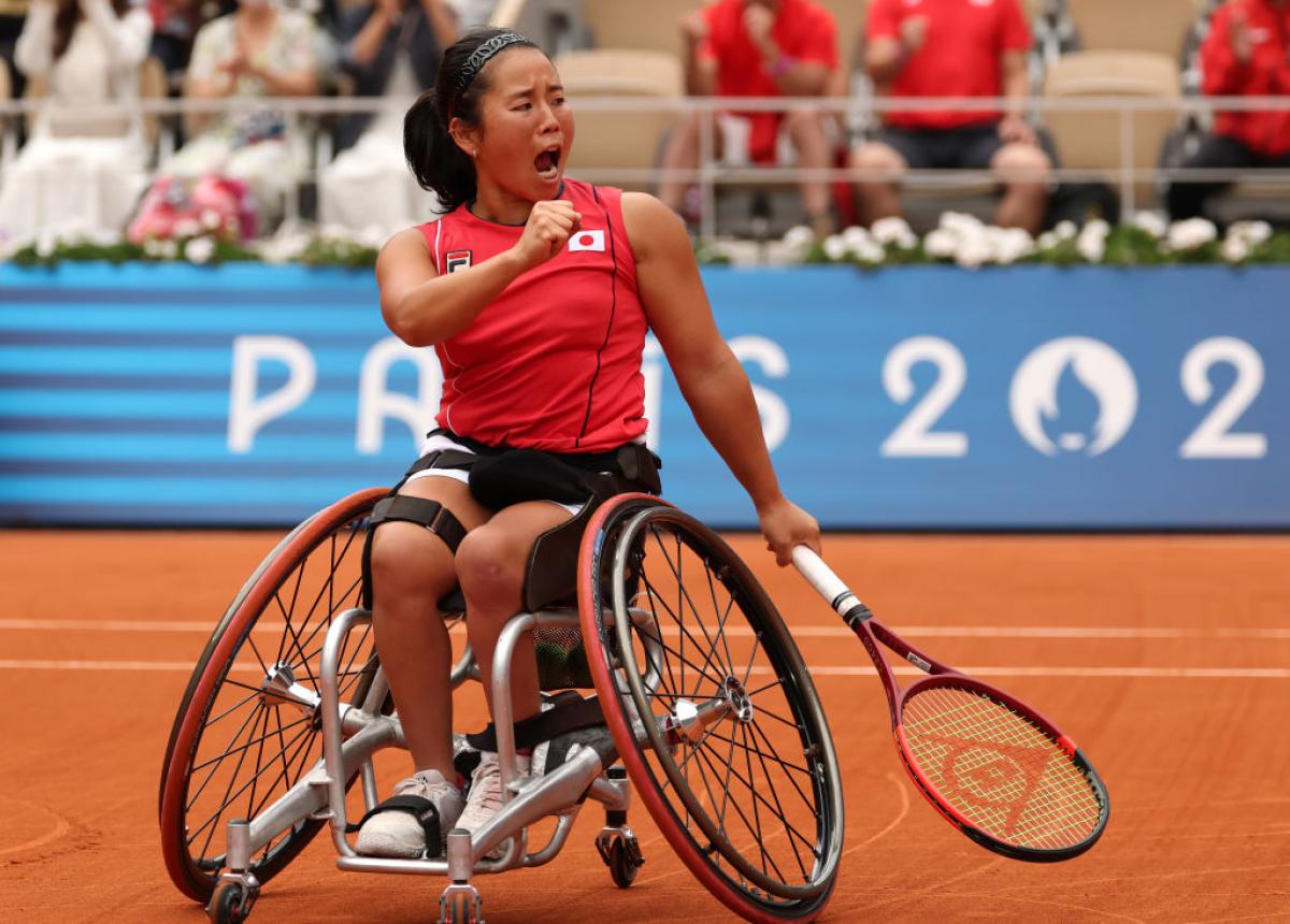 A woman in a wheelchair and a Team Japan shirt holds a tennis racket and yells in celebration