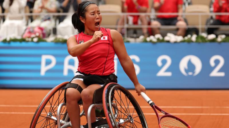 A woman in a wheelchair and a Team Japan shirt holds a tennis racket and yells in celebration