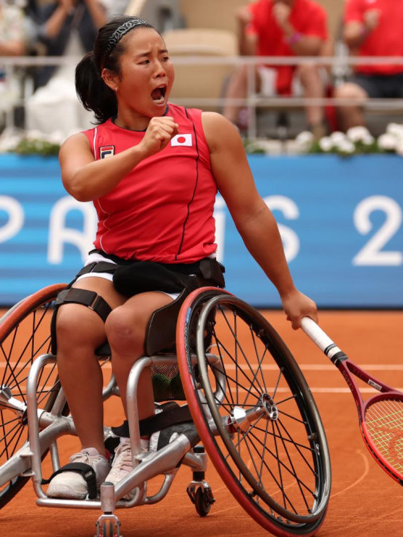 A woman in a wheelchair and a Team Japan shirt holds a tennis racket and yells in celebration