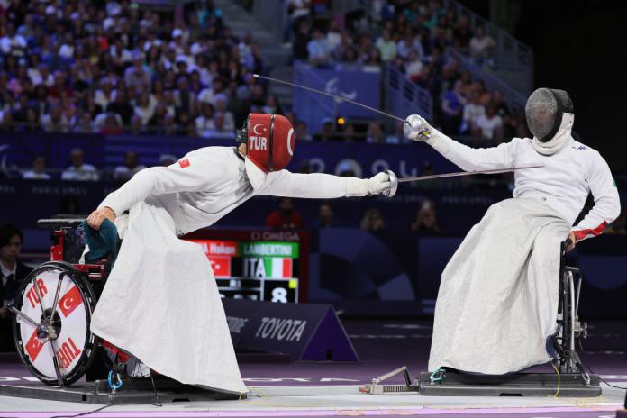 Two people compete in wheelchair fencing. One athlete with a red face shield lunges towards his opponent.
