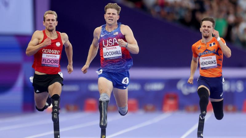 Three men with running blades race against each other, with the man in the middle, in a Team USA uniform, ahead of the other two