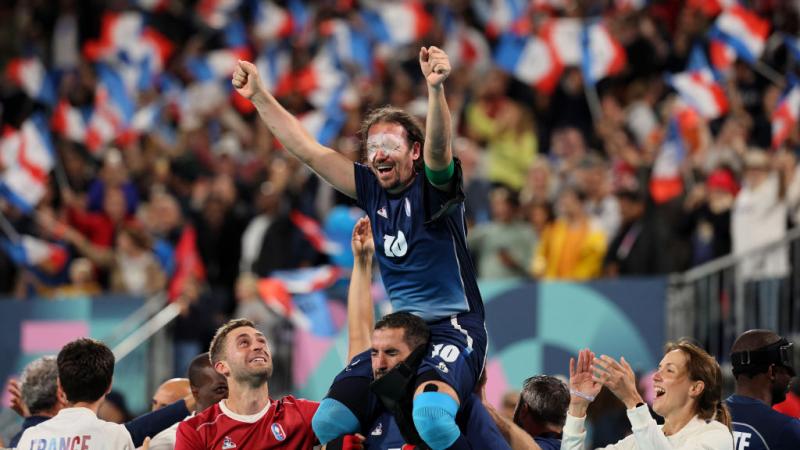 A man in a Team France football uniform with white patches over his eyes is lifted onto the shoulders of one of his teammates, as the crowd in the background waves French flags 