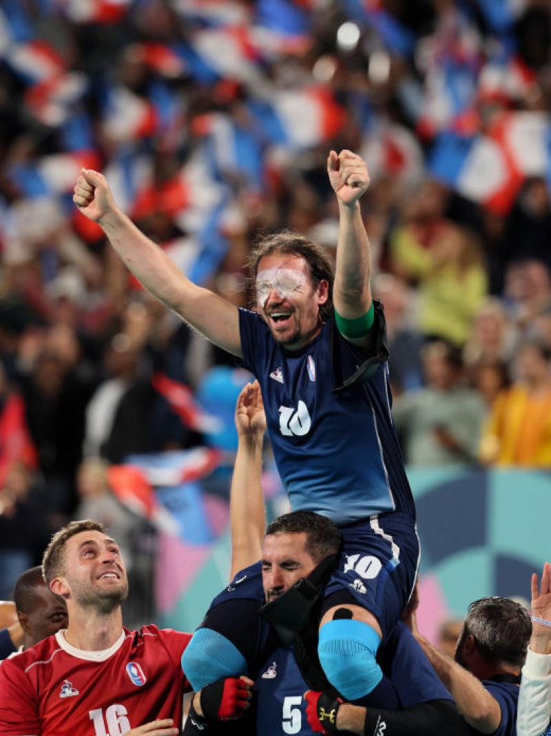 A man in a Team France football uniform with white patches over his eyes is lifted onto the shoulders of one of his teammates, as the crowd in the background waves French flags 