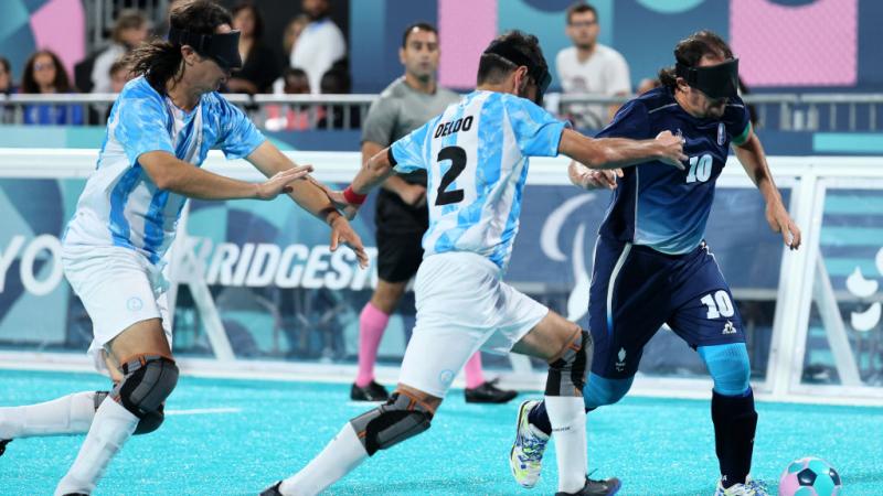 Two blind football players in Argentina uniforms and black eyeshades chase a French player, who has the ball