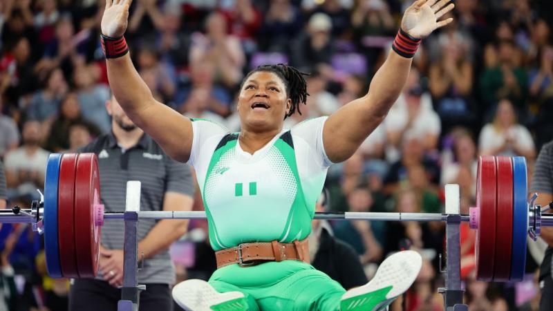 A Para powerlifter in a Team Nigeria uniform sits up on a lifting bench and raises her arms in the air in celebration