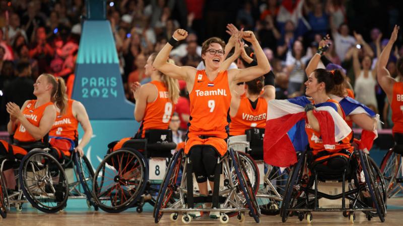 The Dutch women's wheelchair basketball team clap and raise their arms in the air, with one woman in the middle lifting up both arms and smiling