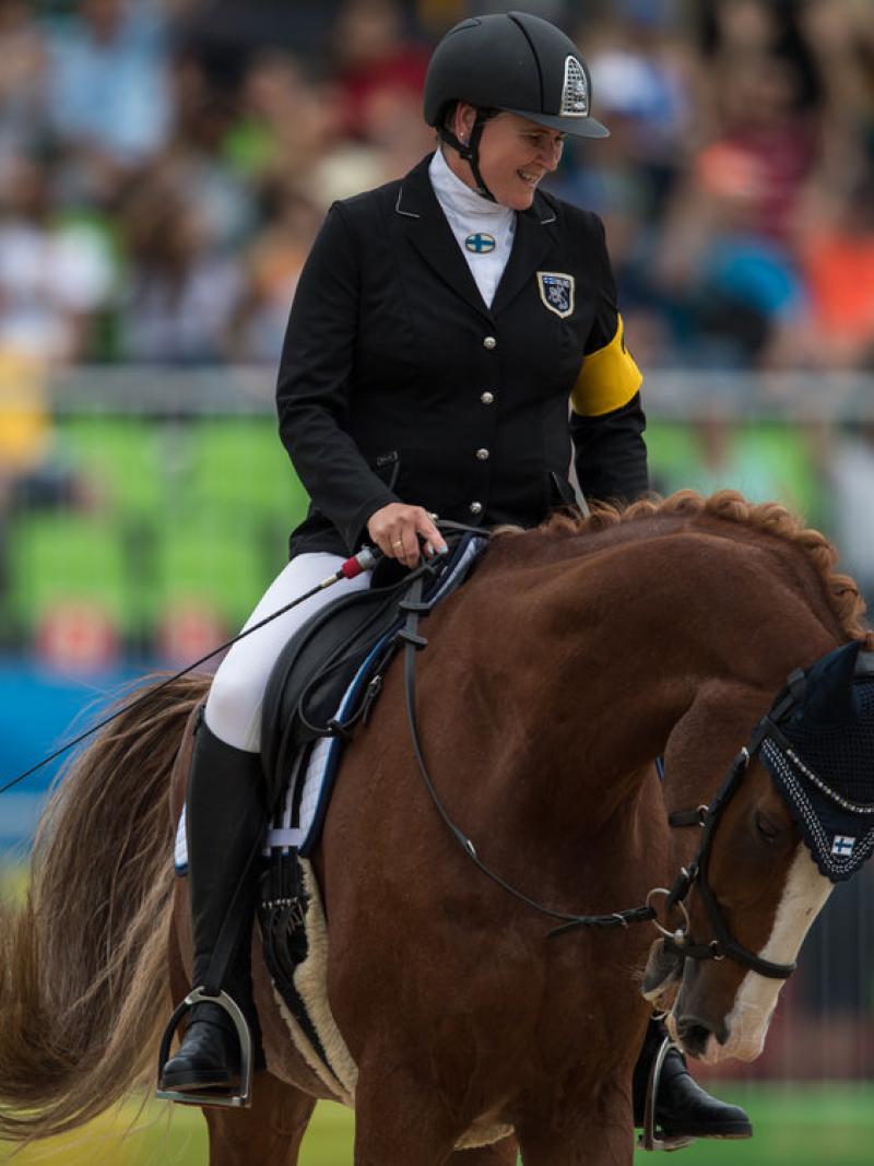 A female Para equestrian rider smiles as she rides a horse during competition.