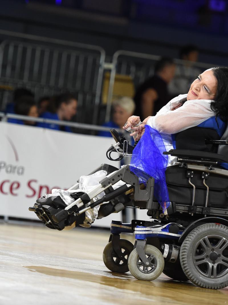 A female dancer in an electric wheelchair smiles and motions with her hands during a dance performance.