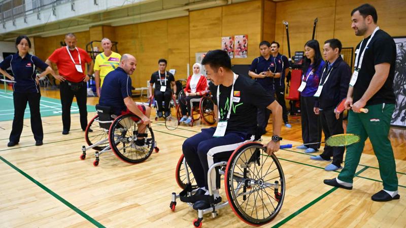 Two men holding badminton racquets are demonstrating on wheelchairs in front of 10 people